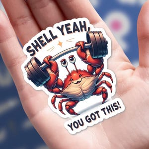 May include: A sticker featuring a cartoon crab lifting weights. The crab is red with a smiling face and holding a barbell with dark gray weights. Text on the sticker reads "SHELL YEAH, YOU GOT THIS!"
