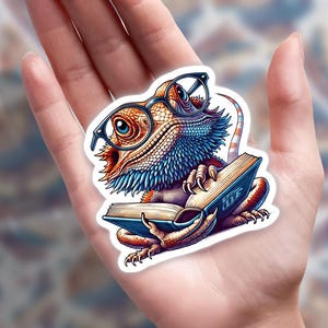 May include: A colorful sticker featuring a cartoon bearded dragon wearing glasses and reading a book. The lizard has blue, orange, and white scales. The book is open, with blue covers and white text. The sticker is held in a hand.