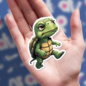 May include: A green cartoon turtle sticker with a grumpy expression. The turtle has a tan shell and is walking with one leg raised. The sticker is held in a hand.