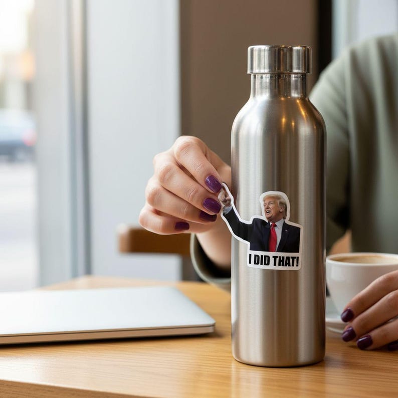 A person is holding a metal water bottle with a sticker of Donald Trump on it, sitting at a table with a laptop and a cup of coffee.