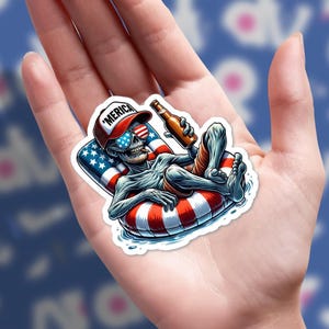 May include: A sticker featuring a cartoon skeleton relaxing on a red and white striped inner tube, holding a beer bottle. The skeleton wears sunglasses, a hat with "'MERICA" on it, and red swim trunks. The inner tube has a section with the American flag.