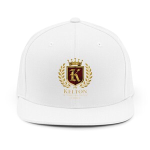 May include: A white baseball cap featuring a centred graphic of a gold crown above a shield with a large gold letter K on a dark red background, surrounded by gold laurel leaves. Below the shield is the text KELTON FAMILY REUNION 2026.