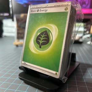 May include: A stack of green Pokemon Basic Energy cards in a plastic sleeve. The card features a green leaf symbol with a white outline. The text on the card reads "Basic Energy" and "ENERGY".