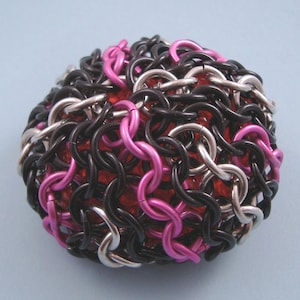 Puede incluir: Un objeto esférico compuesto por anillos metálicos entrelazados en negro, plateado y rosa. Los anillos están dispuestos sobre un núcleo rojo texturizado. La forma general es redonda, con una superficie compleja y texturizada.