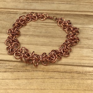 Może przedstawiać: Bransoletka w kolorze miedzi, złożona z wielu małych, połączonych ze sobą pierścieni. Bransoletka ma wygląd łańcucha, z zapięciem. Biżuteria jest prezentowana na jasnej drewnianej powierzchni.