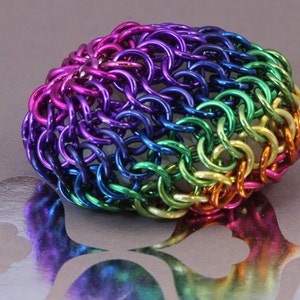 May include: A multi-coloured chainmail bracelet. The bracelet is constructed from small, interlocking metal rings, displaying a gradient of colours such as purple, blue, green, yellow, and red. The jewellery is on a grey surface.