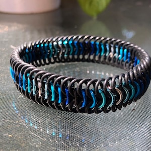 May include: A black chainmail bracelet with alternating sections of blue and silver links. The bracelet is made of metal rings woven together, creating a textured, circular design. The bracelet is shown on a reflective surface.