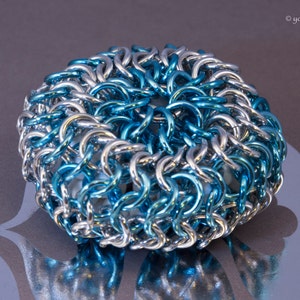 May include: A circular chainmail piece, featuring silver and blue links. The interwoven design creates a textured appearance. It sits on a reflective surface with a subtle pattern. The object is approximately 5 cm in diameter.