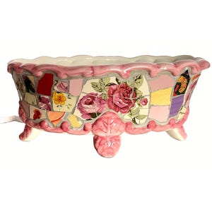 May include: A pink ceramic mosaic planter with a floral design. The planter has a rectangular shape with a scalloped rim and is supported by three small feet. The mosaic features pink, yellow, and red tiles with a rose pattern.