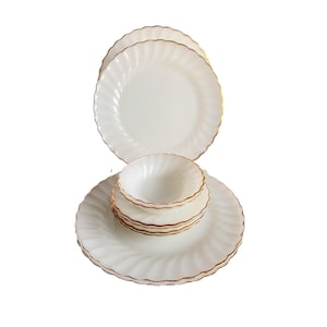 May include: A set of white dinnerware featuring a swirling, fluted pattern along the edges and a thin gold trim. The collection includes two large dinner plates, one stacked vertically and one horizontally, and three smaller bowls stacked on the bottom plate.