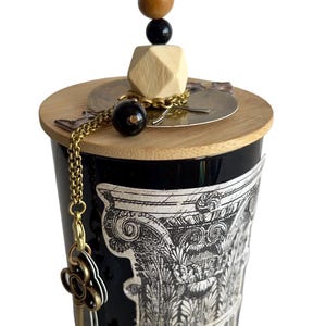 May include: Black candle jar with a wooden lid and decorative key charm. The jar features a detailed black and white illustration. The lid is adorned with a stack of wooden beads and a geometric wooden shape.