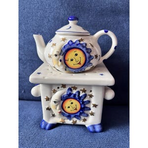 May include: A white ceramic teapot and matching square warmer base featuring hand-painted celestial motifs. The design includes a smiling yellow sun with blue rays and a crescent moon surrounded by small gold stars on both the teapot and base.