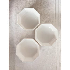 May include: Three white, octagonal ceramic dishes are arranged on a white textured surface. The dishes have a simple, modern design, with a smooth, glossy finish. The plates are empty and ready for use.