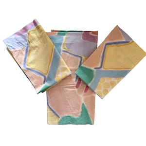 Puede incluir: Tres sábanas de cama dobladas con un colorido patrón abstracto. Las sábanas presentan un diseño con formas amarillas, rosas, azules y verdes. Las sábanas están dispuestas en forma de abanico sobre un fondo blanco.