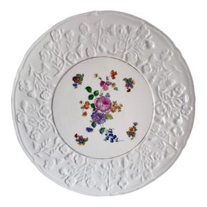 May include: White ceramic plate with a raised floral design around the edge. The centre of the plate features a colourful floral pattern with pink, purple, yellow, and blue flowers.