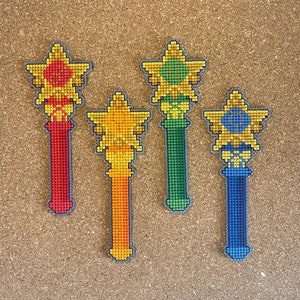 Può includere: Quattro ornamenti ricamati a forma di bacchetta con punte a forma di stella. Le bacchette sono rosse, verdi, arancioni e blu. Ogni bacchetta ha un design diverso al centro della stella.