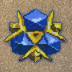 May include: A blue and gold cross-stitch design featuring a stylized figure with a helmet and wings. The figure is surrounded by a golden halo and a geometric pattern.