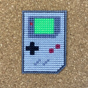May include: A gray and black pixelated cross-stitch of a Game Boy handheld video game console. The screen is green and the buttons are red.
