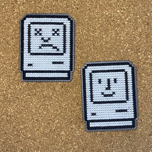 Pode incluir: Dois patches bordados que representam um computador vintage com uma cara feliz e uma cara triste. Os patches são brancos com contornos pretos e estão sobre um fundo de cortiça marrom.