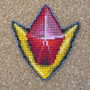 May include: A red and yellow cross-stitch design of a stylized gem with a pointed tip. The gem is surrounded by a yellow border.