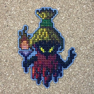 May include: A pixelated embroidered patch depicting a dark-skinned character wearing a yellow and green crown and holding a torch. The character is wearing a red and purple robe.