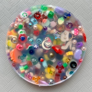 May include: A round, clear resin piece filled with a variety of colorful miniature charms and beads. The charms include a high-heeled shoe, a heart, a star, and a variety of other shapes and colors. The piece is set against a light gray background.