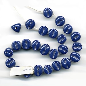 May include: A strand of dark blue glass beads with a textured surface. The beads are shaped like coffee beans and have a small hole in the center. The beads are strung on a white thread and there is a small tag that says "JAPAN".