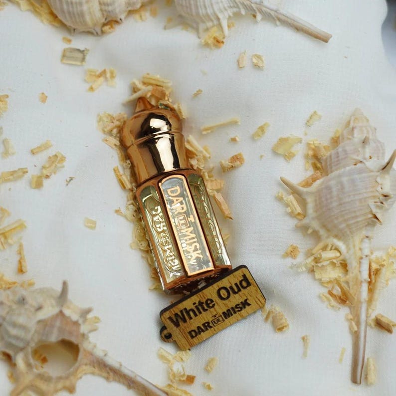 May include: A small, ornate perfume bottle with a gold cap and accents, labeled "White Oud" by DAR@MISK. The bottle is rectangular with gold lettering and is accompanied by a small wooden tag. The background includes seashells and wood shavings.