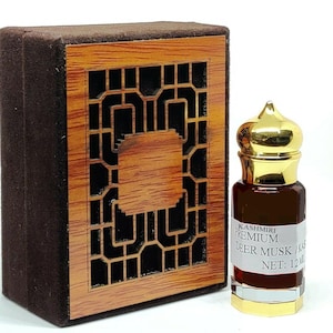 May include: A small, clear glass bottle with a gold cap and a brown liquid inside. The bottle is labeled "Kashmiri Premium Deer Musk / Kas Net: 12 ml". The bottle is sitting on a wooden box with a brown velvet exterior.