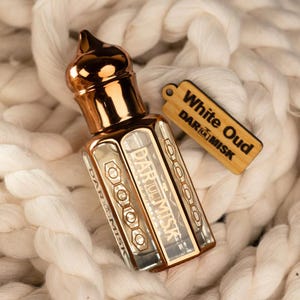 May include: A gold-colored perfume bottle with a decorative top and a rectangular body. The bottle has the text "DAR AL MISK" printed on it. A wooden tag with the text "White Oud" and "DAR AL MISK" hangs next to the bottle. The items rest on a white, textured surface.