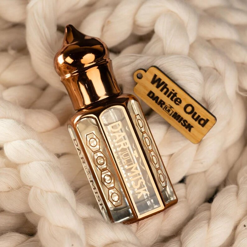May include: A gold-colored perfume bottle with a decorative top and a rectangular body. The bottle has the text "DAR AL MISK" printed on it. A wooden tag with the text "White Oud" and "DAR AL MISK" hangs next to the bottle. The items rest on a white, textured surface.
