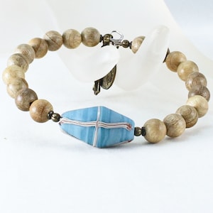May include: A beaded bracelet featuring light brown wooden beads and a central blue, geometric bead with white and red accents. The bracelet is secured with a metal clasp.