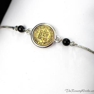 Puede incluir: Pulsera plateada con un medall&oacute;n dorado con detalles intrincados y cuentas negras. El medall&oacute;n tiene un dise&ntilde;o circular con texto y s&iacute;mbolos. La pulsera se muestra sobre un fondo blanco.