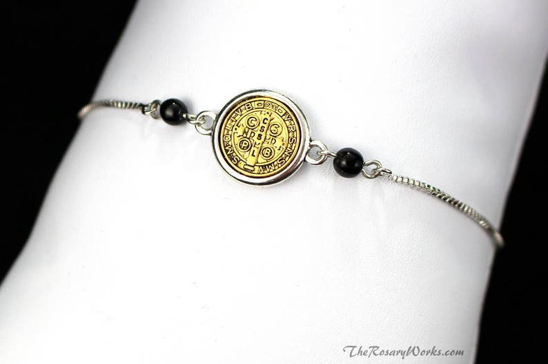 Puede incluir: Pulsera plateada con un medall&oacute;n dorado con detalles intrincados y cuentas negras. El medall&oacute;n tiene un dise&ntilde;o circular con texto y s&iacute;mbolos. La pulsera se muestra sobre un fondo blanco.