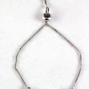 Puede incluir: Pulsera ajustable plateada con un medall&oacute;n religioso dorado y cuentas negras. La pulsera tiene un cierre deslizante para un ajuste personalizado. El texto "TheRosaryWorks.com" es visible en la parte inferior.