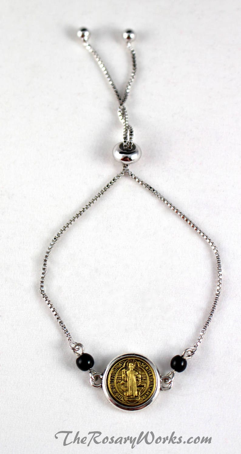 Puede incluir: Pulsera ajustable plateada con un medall&oacute;n religioso dorado y cuentas negras. La pulsera tiene un cierre deslizante para un ajuste personalizado. El texto "TheRosaryWorks.com" es visible en la parte inferior.