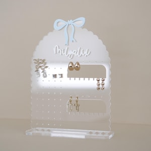 May include: Clear acrylic earring holder with a scalloped edge and a blue bow on top. The holder has a nameplate that says "Mygalie" and multiple rows of holes for earrings.