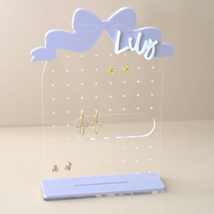 May include: A clear acrylic earring holder with a purple bow and the name "Lily" on top. The holder has multiple holes for earrings and a small shelf at the bottom.