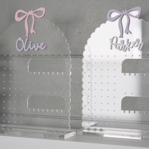 May include: Two clear acrylic earring holders with scalloped edges. The holder on the left has a pink bow and the name "Olive" written on it. The holder on the right has a purple bow and the name "Parker" written on it.