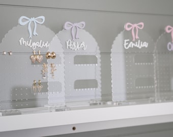 May include: Four clear acrylic earring holders with a scalloped edge and a bow on top. Each holder has a name written on it:  'Mygalia', 'Parker', 'Emilia', and 'Olivia'.