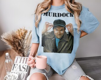 May include: A light blue t-shirt with a graphic print of a man wearing a hat and a leather jacket. The text "MURDOCK" is printed in black on the shirt.