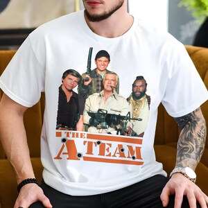 Pode incluir: Camiseta branca com uma estampa gráfica dos personagens do programa de televisão "A-Team". Os personagens estão em pé em frente a uma faixa vermelha e laranja com as palavras "A-Team" impressas nela.
