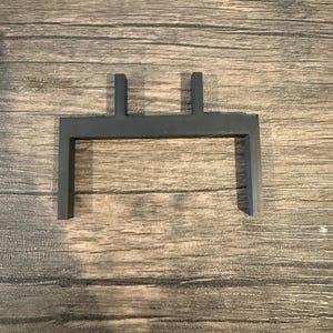 Könnte beinhalten: Eine dunkelgraue, rechteckige Metallhalterung mit zwei vertikalen Pfosten. Die Halterung hat ein einfaches, geometrisches Design und dient wahrscheinlich zur Unterstützung oder Montage. Das Objekt befindet sich auf einer Holzoberfläche.