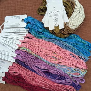 May include: A collection of Gloriana hand-dyed silk embroidery threads in various colors including blue, pink, purple, red, brown, and cream. Each bundle of thread is attached to a white paper tag with printed product information and branding.