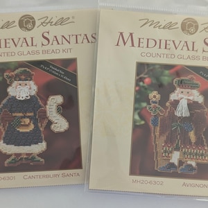 May include: Two Mill Hill Medieval Santas counted glass bead kits. One kit features a Santa in a dark blue and brown outfit holding a scroll. The other kit shows a Santa in a brown robe with a staff. Each kit is 7 x 4.5 inches.
