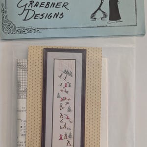 May include: A cross-stitch kit with a framed winter scene depicting skiers and trees. The kit contains a pattern, threads, and fabric. The packaging displays the text "DIANE GRAEBNER DESIGNS". The kit is sealed in a clear plastic bag.