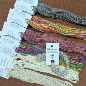 Puede incluir: Una colección de hilos de bordar de seda de 12 hebras teñidos a mano Gloriana en varios colores, incluyendo gris, una mezcla multicolor de rosa, amarillo y verde, rosa viejo y crema. Cada madeja está unida a una etiqueta de papel blanco.