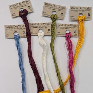 May include: A collection of colorful embroidery floss skeins from Weeks Dye Works. The skeins are in various colors, including blue, burgundy, green, white, pink, and yellow. Each skein is labeled with a color name and number.