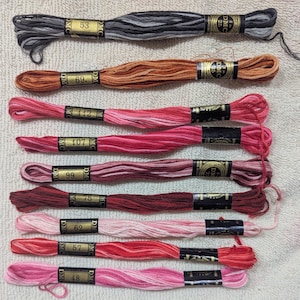May include: A collection of DMC embroidery floss in various colors, including shades of gray, brown, and red. Each skein is labeled with a number and the DMC brand name. The floss is neatly arranged, showcasing the range of hues available for crafting projects.