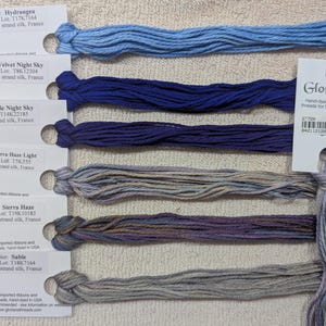 May include: A selection of hand-dyed silk threads in various shades of blue, purple, and gray. Each skein is labeled with color names like "Hydrangea" and "Velvet Night Sky." The threads are 6 yards long.